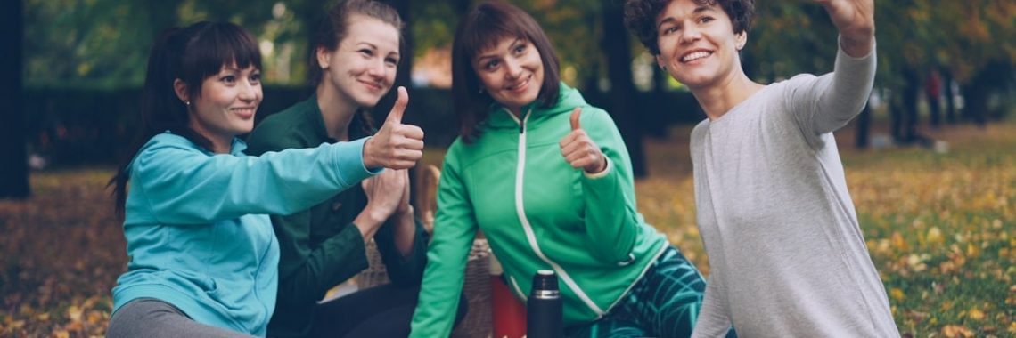 Attractive young ladies yogini are taking selfie using smartphone during picnic in park in autumn. Girls are posing and smiling having fun sitting together on mats.